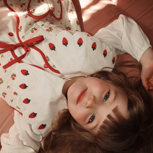 Young girl wearing a white dress with red strawberry pattern lying on a pink surface.